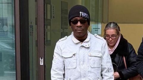 Henry Morgan, a man wearing a black woollen hat, pale denim jacket and sunglasses, pictured close-up exiting a set of glass doors.