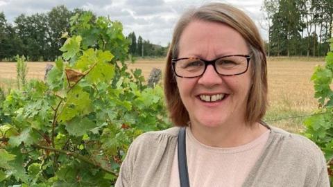 Woman smiles at the camera,  wearing glasses with green fields in the background