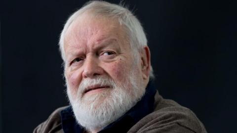 Michael Longley has short white hair and beard and is wearing a brown cardigan over a dark blue shirt.