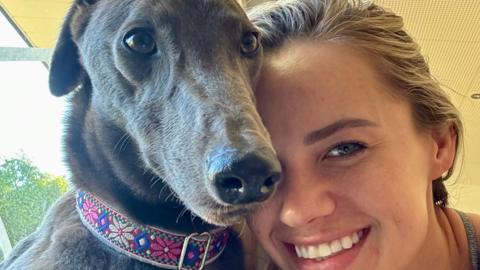 Kiki the grey greyhound with brown eyes. She is wearing a colourful collar. She is with Natalie Angst who is smiling and has blonde hair tied back. They are indoors and a tree is visible through a window.