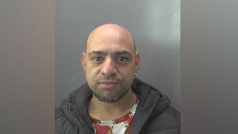 Mohammed Fiaz is looking at the camera in a police custody photo. He is mostly balding with a beard and moustache. He has an earring in his left ear. He is wearing a red patterned top and a black jacket
