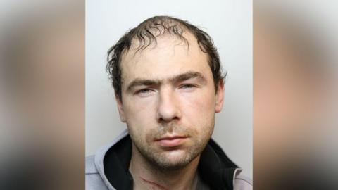 Mugshot of Joshua McMenamin, he has thin dark hair, a scratch on his neck and wears a grey hoodie