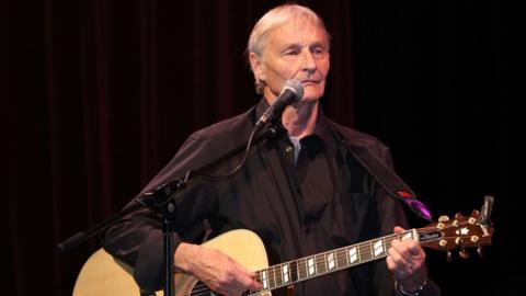 Len Garry is pictured on stage with grey hair and wearing a black shirt and playing an acoustic guitar