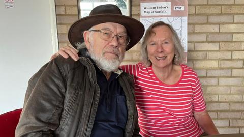 John and Linda Truscott sit on chairs around a table based in front of an indoor brick wall. John wears a brown hat, leather jacket and blue top. He has a beard and is wearing glasses while smiling at the camera. Linda has her arm around his shoulders and is wearing a red and white striped top. She has shoulder length, grey hair and is also smiling.