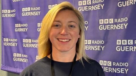 Charlie Cox is smiling at the camera as she stands next to a BBC Radio Guernsey branded banner. She has shoulder-length blonde hair and blue eyes., She is wearing a black top and a lanyard.