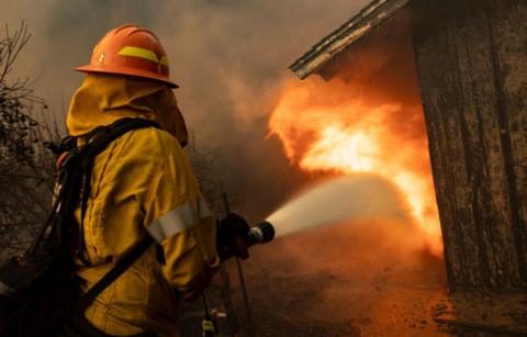 A firefighter in yellow protective gear uses a hose to control a blaze on a burning wooden shed in California