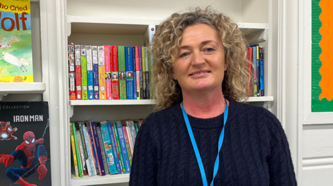 Mel Medlock stands in front of a mini library full of brightly coloured books on white shelves. She has blonde curly hair and wears a navy jumper.