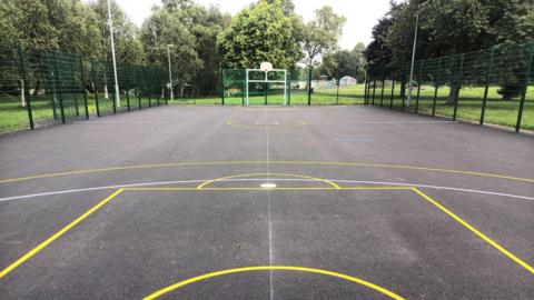 The refurbished multi use games area. The playing pitch is within the park, surrounded by grass and trees. It has goal posts and basket ball hoops and lines of different colours on the ground to show the pitch areas for different games.