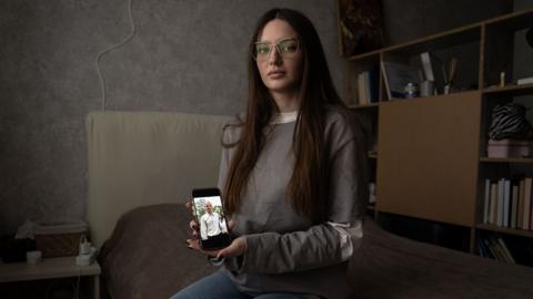 Julia Hripun sitting in her bedroom with a picture of her captive father on her iPhone