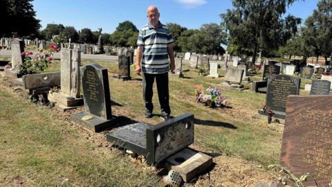 A man wearing a striped t-shirt and dark jeans stands in a cemetery. He is next to a black gravestone which has been laid flat on the ground. Other gravestones and floral tributes are visible in the background.