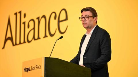 Andy Burnham has short dark hair and stands at a lectern. He is wearing a suit without a tie, and glasses. Behind him is a large yellow screen with 'Alliance' written on it. On the lectern 'Hope. Not Fear.' is written.