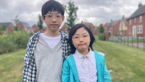 Elias, a boy wearing a grey and white shirt with a blue checked overshirt, standing next to his sister Ariel, wearing a white shirt and light blue jacket.