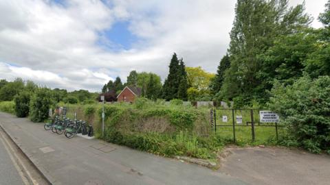 Google street view of empty overgrown land with high bushes connecting to a metal gate. There are bikes line up in a bike rack against the bushes