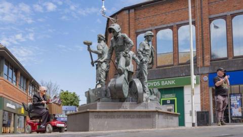 The Spirit of Barrow sculpture, which celebrates the port town's history of shipbuilding. It features three men in work overalls and protective headwear carrying hammers and rope. A woman is riding past on a mobility scooter, while a man standing to the right-hand side of the artwork is talking on a mobile phone. Branches of Lloyds Bank and McDonald's can also be seen in the background.