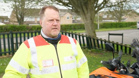 A man stood in a grassy garden with a fence behind him and a road in the distance. He has short brown hair and a short beard. He has a florescent jacket on. His orange motorbike is next to him.