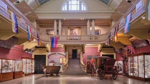 Royal Cornwall Museum interior. Stairs in the centre of the museum lead up to a balcony which over looks the inside of the building. It is pink and yellow. Display cupboards line the walls.