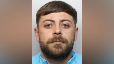 Mugshot of Jordan Woolmer. HE is wearing a light blue jacket and has short brown hair and beard.