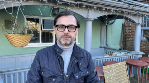 A man in a blue jacket and glasses standing in front of a pub 