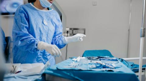 A woman in a clinical gown, mask and gloves prepares surgical instruments on a trolley covered in a blue cloth in an operating theatre.