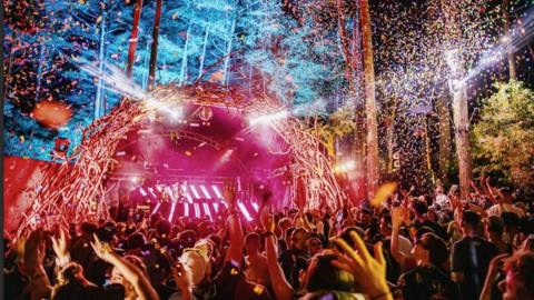 General image of a crowd partying with their hands in the air in front of a brightly lit stage in pinks and blues