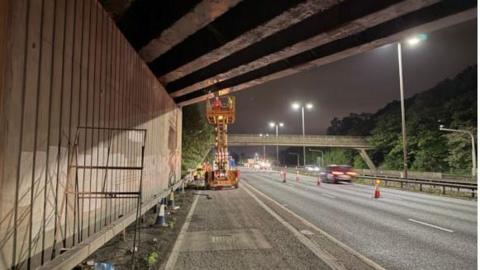 Two workers on a cherry picker in the distance on a hard shoulder under a bridge. A three-lane motorway can be seen next to them with cones marking off two lanes. Blurred cars drive past.