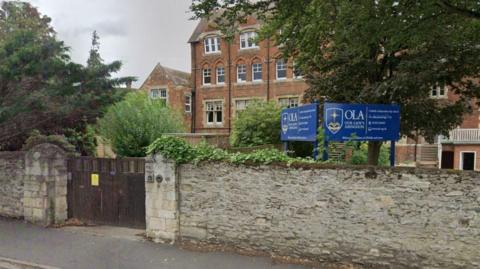 A Google Maps closeup of the front of red brick private school. A wall sits in front of the building with a school gate to the left, and a sign for the school rests just above the wall. 