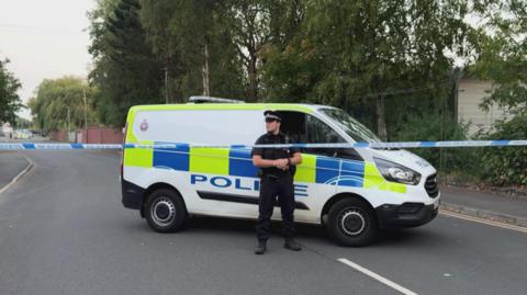A police officer in black uniform stands next to a liveried van, with a line of blue and white police tape in the foreground