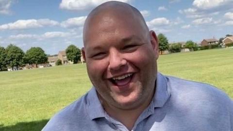 Gary Stanley: the head and shoulders of a smiling man who has close-cropped hair. He is wearing a pale blue shirt, which has a collar and is unbuttoned at the neck. An area of green grass - plus green-leaved trees and brown buildings - are in the background.
