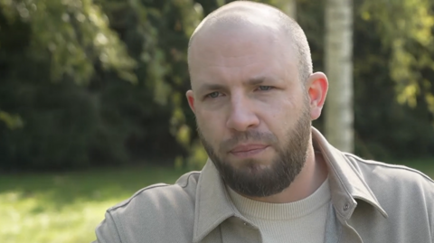 A bald man with brown facial hair. He is looking off to the left of the image and is wearing a cream t-shirt and cream jacket. Behind him are blurred trees and grass