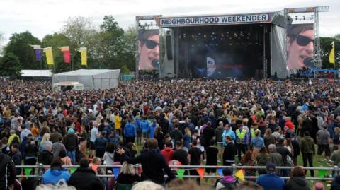 The Neighbourhood weekender stage with Gerry Cinnamon performing. Above the stage a white sign with black writing reads Neighbourhood Weekender. Each side of the stage there are two large screens showing the performer Gerry Cinnamon singing into the microphone. In front of the stage, a large crowd is gathered watching.