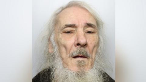 An elderly man with long white hair and beard. He is standing against a grey background wearing a black top.