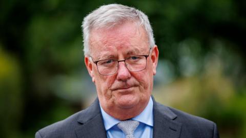 Mike Nesbitt is outside. He's wearing glasses and a grey suit. His tie has a maze design. His shirt is light blue. The background is out of focus.