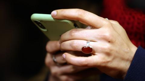 A close-up of a woman's hands holding a smartphone. She has a ruby-coloured ring on the third finger of her left hand.