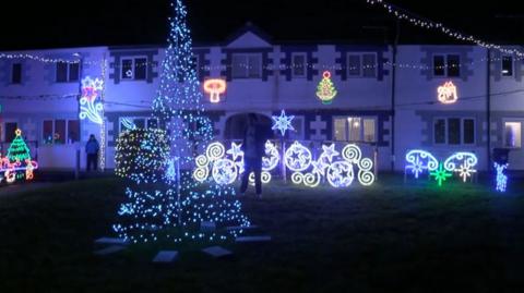 A Christmas tree is lit up in front of a white house with other Christmas decorations.