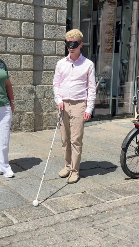 Man in a pink shirt and cream trousers with a black blindfold and holding a white cane