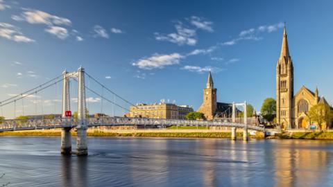 A bridge over a calm river with Victorian building on the far bank, under a largely blue sky