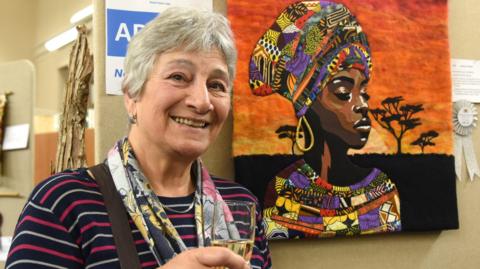 Karen Wright with short grey hair smiling and holding a glass of champagne next to her picture of an African woman made of fabric built up to depict shading on her face and bright colours on her clothes