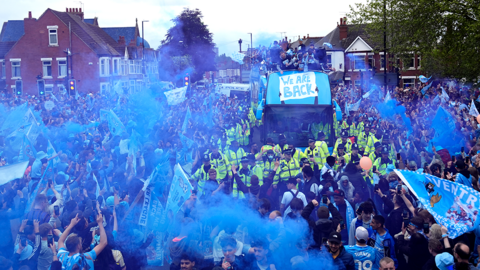 Thousands of people crowd around a bus surrounded on all sides by police officers in high-vis jackets. many of the people are carrying flares giving off lots of blue smoke. Many are also carrying light blue Coventry City flags. At the centre is the open top bus with the team at the top. At thr front is a sign, reading: "We are back"