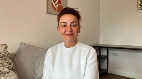 Kelly Slack is smiling and looking at the camera. She is wearing a plain white t-shirt and multiple earrings. Behind her are large grey cushions and a white wall with a framed print of a plant on it. 