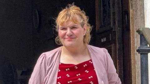 Katie Wallis walking out of court building wearing red top with white bows and pink blazer over it. She has long curly blonde hair up in a pony tail.