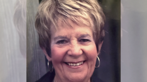 A late middle-aged woman smiling at the camera with partially dyed hair and wearing hooped earrings.