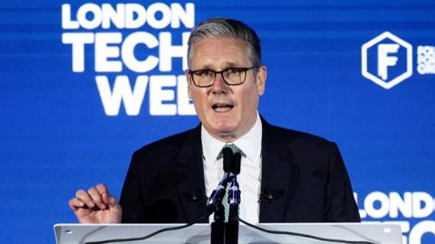 Starmer gestures as he speaks at a lectern in front of a blue background reading "london tech week".