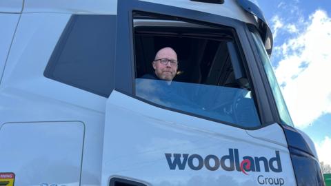 A man wearing glasses and a black jumper sat inside of the cab of a large white lorry, looking out of the window on the driver's side with the window wound down. 