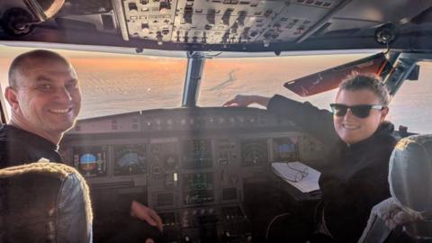A man and a woman sit in the cockpit of a plane with the dashboard visible and snow stretching out on the Earth through the window