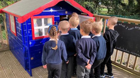School aged children in front of a blue and red Wendy house.