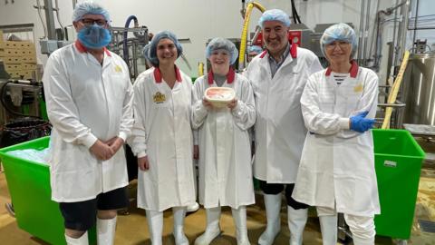 Four adults standing in white coats in a dairy, with a boy in the middle holding a tub of ice-cream