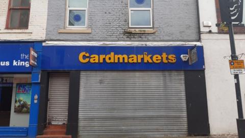 The shop on Blandford Street. It is a two-storey building with a tatty shutter over the bottom floor and a yellow sign reading "Cardmarkets". There are two windows upstairs.