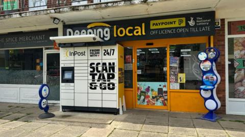 The outside of a small shop with bright orange paint, a National Lottery stand and the words Deeping Local above the window. There is a unit of InPost lockers standing outside.