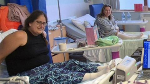 Two women in hospital beds smiling for the camera. On the left is a woman with brown hair in a plait and glasses, who is leaning forward on a bed, wearing a black vest and blue patterned trousers. Next to her is another woman in a grey jumper and knee high socks with long blonde hair leaning forward and smiling too.