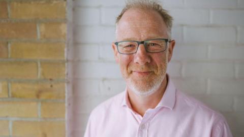 Ben wearing a pastel pink shirt with ginger and grey stubble wearing blue rimmed glasses smiling at the camera. He is standing in front of a brick wall. Some of it has been painted white.